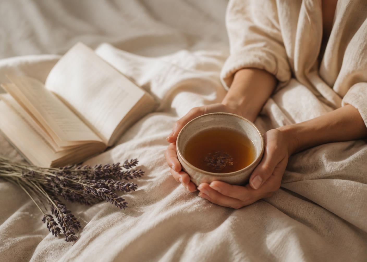 Mani che tengono una tazza di ceramica con infuso, accanto a un libro aperto e lavanda secca su lino